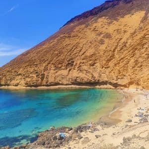 Una cala de playa arenosa con aguas cristalinas de color turquesa al pie de un acantilado empinado de color ocre bajo un cielo azul brillante.