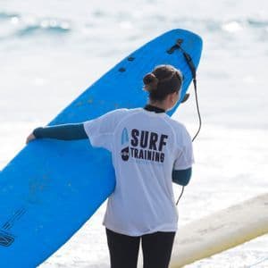Una mujer en traje de neopreno, vista de espaldas, sostiene una tabla de surf azul grande mientras está de pie en el agua poco profunda del océano.