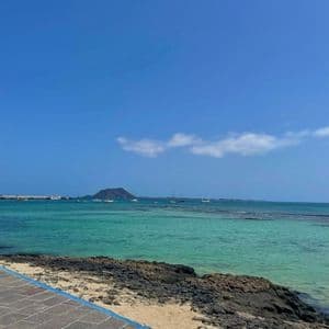 Un paseo de piedra con vistas a una bahía de aguas turquesas, donde veleros están anclados con una gran isla al fondo bajo un cielo azul.