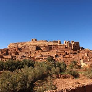 A traditional mud-brick village built on a hillside under a clear blue sky, with trees and fields in the foreground.