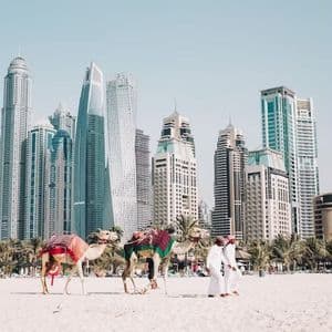 Deux hommes en robes traditionnelles marchent avec deux chameaux sellés sur une plage de sable, avec une ligne d'horizon de ville moderne en arrière-plan.