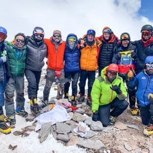 A WeRoad group trip of people in mountaineering gear posing for a photo on a snowy, rocky mountain summit.