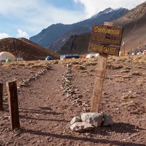 A wooden sign for Confluencia camp sits on a rocky path leading to a high-altitude campsite with tents at the base of mountains.