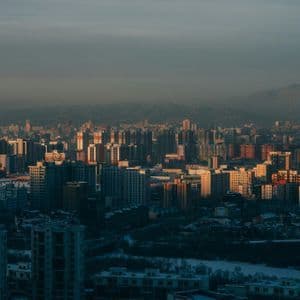 A dense cityscape with numerous buildings illuminated by warm, low sunlight, with hazy mountains in the background.