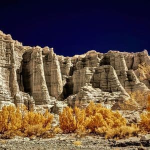 Jagged, light-colored rock formations rise above yellow trees on a rocky terrain under a deep blue sky.