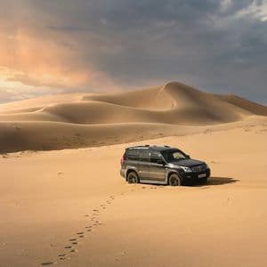 A dark SUV is parked on a desert sand dune with footprints leading away from it under a cloudy sunset sky.