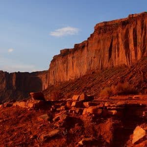 Large red rock formations glowing in the warm sunset light, with a rocky foreground and a blue sky.