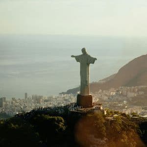 A large statue with outstretched arms stands on a verdant mountain peak, overlooking a coastal city with a crowd of people at its base.