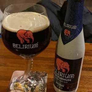 Un bicchiere di birra scura Delirium Nocturnum con una schiuma densa, posto accanto alla sua bottiglia abbinata su un tavolo di legno.