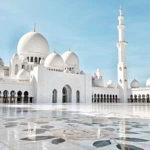 Le cupole bianche e il minareto di una moschea si innalzano sopra un cortile in marmo lucido con intarsi floreali sotto un cielo azzurro limpido.