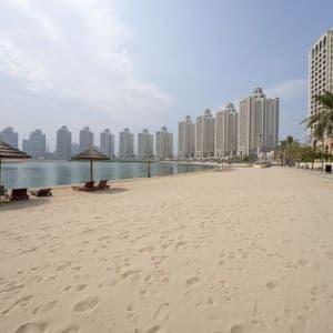 Un'ampia spiaggia sabbiosa con ombrelloni e sedie a sdraio di fronte a un moderno skyline cittadino oltre l'acqua.
