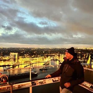 Un hombre con gorro y chaqueta está en un mirador, contemplando un paisaje urbano iluminado y un río al anochecer.