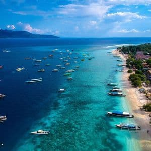 Una vista aérea de muchos barcos flotando en agua turquesa a lo largo de una playa de arena, con montañas en el horizonte bajo un cielo parcialmente nublado.