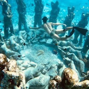 Una persona con máscara y aletas hace snorkel sobre un parque de esculturas submarinas en el arenoso fondo marino.