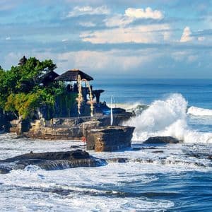 Un templo se asienta en una pequeña isla rocosa cubierta de árboles, con grandes olas del océano rompiendo contra la orilla bajo un cielo parcialmente nublado.
