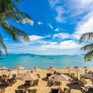 Filas de tumbonas y sombrillas vacías bordean una playa de arena, frente a un mar turquesa con barcos bajo un cielo azul y parcialmente nublado.