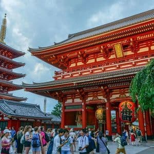Una multitud de personas se reúne en un patio frente a una gran puerta roja de templo y una pagoda de cinco pisos bajo un cielo nublado.