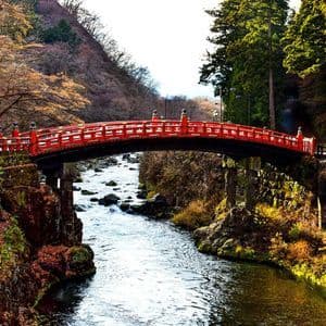 Un puente rojo y ornamentado con arcos cruza un río rocoso que fluye por un valle boscoso con follaje de otoño.