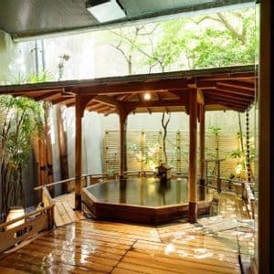 Un onsen tradicional de madera en un patio exterior, con una glorieta, tarima mojada y plantas de bambú.