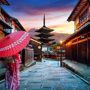 Una persona con un kimono sostiene un paraguas rojo en una calle empedrada bordeada de casas tradicionales y una pagoda al atardecer.