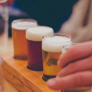 Primer plano de una degustación de cervezas con cuatro variedades diferentes en vasos sobre una bandeja de madera, y una mano alcanzando uno.