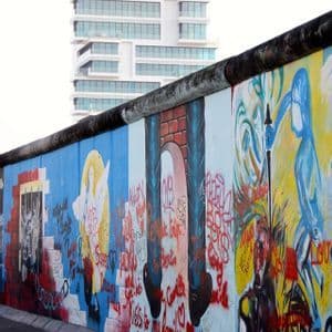 Una larga pared de hormigón cubierta de coloridos murales y grafitis, con un edificio moderno visible al fondo.