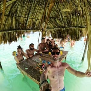 Un gruppo WeRoad scatta un selfie, seduto a un tavolo di legno immerso nell'acqua turchese, sotto una capanna dal tetto di paglia.