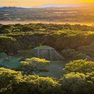 Ein alter Steintempel steht auf einer Dschungellichtung, beleuchtet von goldenem Licht einer untergehenden Sonne über fernen Bergen.