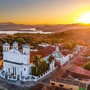 Eine Luftaufnahme einer weißen Kolonialkirche und einer Stadt mit roten Ziegeldächern, mit Blick auf einen See und Berge bei Sonnenuntergang.