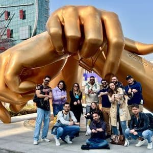 Un viaggio di gruppo WeRoad in posa davanti a una scultura gigante dorata di mani incrociate in una piazza urbana.