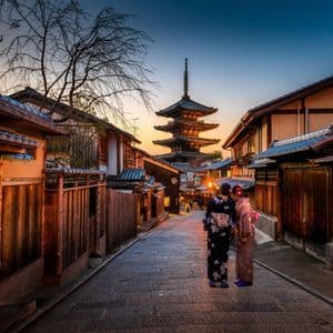 Due donne in kimono si trovano su una strada di ciottoli, osservando una pagoda a più livelli al tramonto, fiancheggiata da case tradizionali.