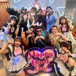 Un gruppo WeRoad in viaggio posa con donne in abiti da cameriera in un caffè a tema, reggendo un grande cartello 'Buon Compleanno'.