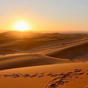 Il sole cala sulle sinuose dune di sabbia del deserto, con impronte e tracce di pneumatici visibili nella sabbia dorata.