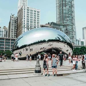 A crowd of people gathers around a large, reflective, bean-shaped sculpture in a city plaza, with skyscrapers in the background.