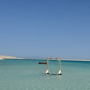 Un'altalena di legno si erge in acque poco profonde e limpide di colore turchese sotto un cielo di un azzurro intenso, con una costa sabbiosa in lontananza.