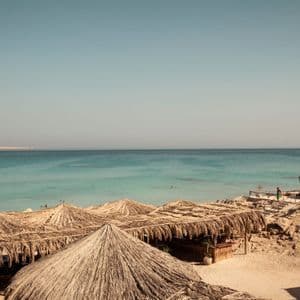 Una vista dall'alto di una spiaggia sabbiosa con ombrelloni dal tetto di paglia, che si affaccia su due barche ormeggiate in acqua turchese sotto un cielo sereno.