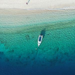 Vista aerea di una barca ancorata in acqua turchese accanto a una spiaggia di sabbia bianca con una persona che cammina lungo la riva.