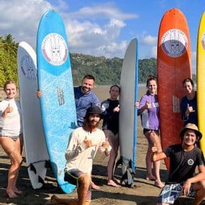 Eine WeRoad Gruppenreise posiert mit bunten Surfbrettern an einem Sandstrand unter blauem Himmel.