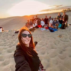 Une femme prend un selfie avec un groupe WeRoad posant ensemble sur des dunes de sable au coucher du soleil doré.