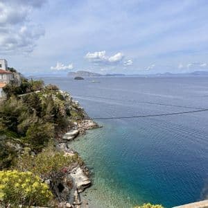 Una vista dall'alto di una costa rocciosa con alberi verdi, una piccola caletta con acqua turchese e isole lontane sotto un cielo nuvoloso.