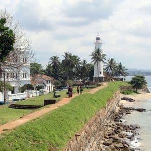 Una vista panorámica de un faro blanco y palmeras en una costa verde con un muro de piedra y el océano.