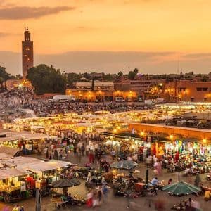 Vista dall'alto di una vivace piazza del mercato cittadino al tramonto, gremita di folla e bancarelle luminose, con una torre sullo sfondo.