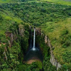 Luftaufnahme eines Wasserfalls, der von einer Klippe in ein Becken in einer üppig grünen Schlucht stürzt, umgeben von sanften Hügeln.