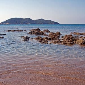 Ein felsiges Ufer mit klarem, flachem Wasser, das an einen Sandstrand schwappt, mit einer grünen Insel am Horizont.