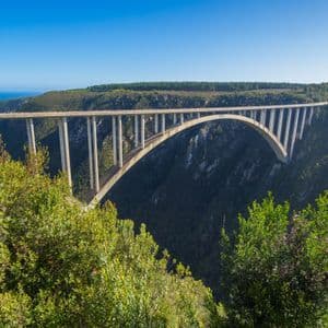 Eine Betonbogenbrücke überspannt eine tiefe, bewaldete Schlucht mit dem Ozean in der Ferne unter klarem Himmel.
