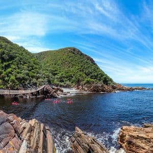 Eine WeRoad Gruppenreise paddelt in rosa Kajaks auf einem dunklen Fluss unter einer Hängebrücke, wo grüne Hügel auf das Meer treffen.