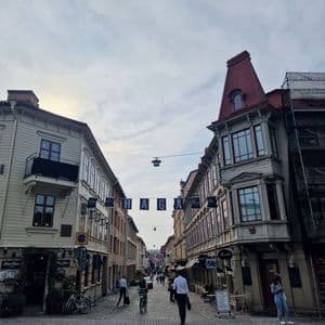 Menschen gehen und radeln auf einer Kopfsteinpflasterstraße, die von traditionellen, mehrstöckigen Gebäuden unter einem bewölkten Himmel gesäumt ist.