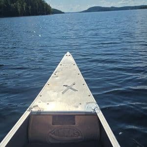 Eine Ich-Perspektive vom Bug eines Aluminiumkanus, das über einen großen, funkelnden See auf ein bewaldetes Ufer unter blauem Himmel gleitet.