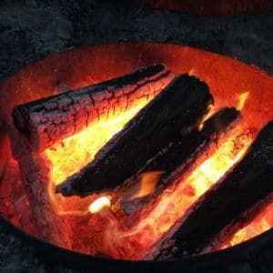 Mehrere verkohlte Holzscheite brennen mit hellorangen Flammen und glühenden Kohlen in einer Metall-Feuerstelle bei Nacht.