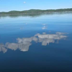 Ein ruhiger See spiegelt den blauen Himmel und die weißen Wolken darüber perfekt wider, mit einem bewaldeten Ufer im Hintergrund.